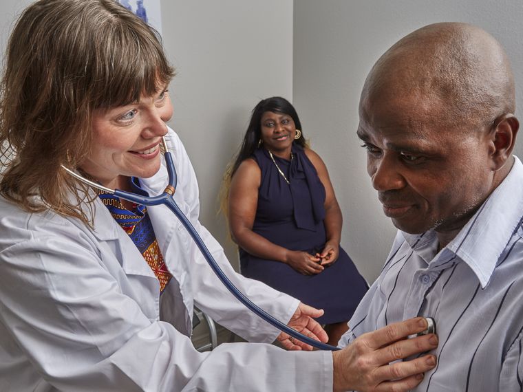 A clinic provider listens to a patient's heart with a stethoscope while a second patient waits nearby.